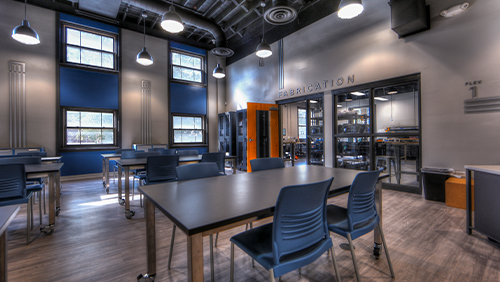 The Flex 1 space. A Room with high, black colored ceilings, and a blue accent wall with 4 large windows. It is filled with 6 long tables with grey tops and wooden legs on wheels. Each table has 4 blue chairs at it. You can see the glass widows and entrance to the Fabrication room in the background. 