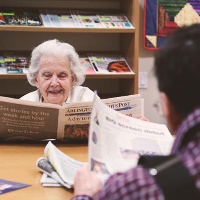 A smiling woman and man reading newspapers in the senior center reading room